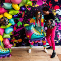 Two people in colorful clothes smile and pose with a 27" SuperShape 90s Party Inline Roller Skate Foil Balloon by Anagram/MD and SO FLY balloons in front of a neon splatter-paint backdrop, standing on a wooden floor.