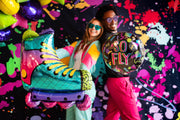 Two people in colorful retro outfits and sunglasses pose in front of a vibrant splatter-paint backdrop. One holds an Anagram/MD 27" SuperShape 90s Party Inline Roller Skate Foil Balloon; the other holds a round “SO FLY” balloon.