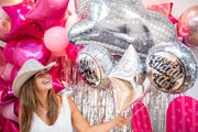 A smiling woman in a white dress and cowboy hat holds the Anagram/MD Bouquet Disco Western Foil Balloon, standing before a festive backdrop of pink and silver balloons and metallic streamers.