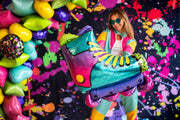 A woman in a colorful outfit and sunglasses holds the Anagram/MD 27" SuperShape 90s Party Inline Roller Skate Foil Balloon in front of a splatter-paint backdrop with bright star-shaped balloons, creating a playful, festive scene.