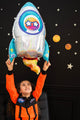 A child in an orange astronaut suit smiles while holding an Anagram/MD 29" Holographic SuperShape Outer Space Foil Balloon. Behind them, a black wall decorated with stars and planets creates a fun space-themed backdrop.