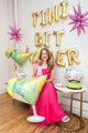 A smiling woman in a bright pink dress sits beside a birthday cake and a 36" SuperShape Cocktail Surprise Foil Balloon by Anagram/MD, with gold "TINI BIT OLDER" letter balloons and pink star decorations in the background.
