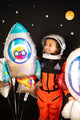 A young child beams while holding the 29" Holographic SuperShape Outer Space Foil Balloon by Anagram/MD—a shiny, rocket-shaped balloon—among colorful, space-themed decorations against a starry black background.