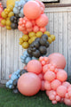 A vibrant balloon garland featuring the Tuftex 36″ Poppy Tuftex Latex Balloons Designer Balloons (2 per bag) in pink, yellow, blue, and brown, arranged against a weathered white wooden wall on green grass.