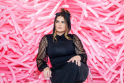 A woman with long wavy hair and a black headband sits on a clear chair, in front of a wall decorated with TT-260's Baby Pink Tuftex Brand Latex Balloons for animal twisting by Tuftex. She wears a black sheer-sleeve top and black pants.