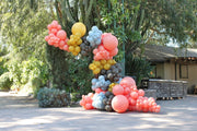 A vibrant balloon sculpture made with Tuftex 5 Inch Latex Balloons in the Designer Poppy collection stands outdoors near greenery and a cactus, set against a rustic building.