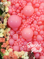 A display featuring a wall of Kalisan 12″ Queen Pink heart-shaped latex balloons in varying sizes, accented with clusters of colorful flowers along the left and bottom edges. The words “Queen Pink” appear on the image, with a dotted white arrow directing attention from the floral arrangements to the Kalisan balloons.